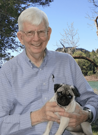 Smiling man in glasses holds a small pug on his lap, sitting outdoors with trees and a river in the background.
